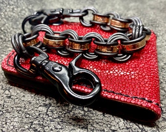 May include: A black metal chain bracelet with gold-coloured accents rests on a red leather wallet. The bracelet has a lobster clasp and is connected to the wallet with a black metal clasp.