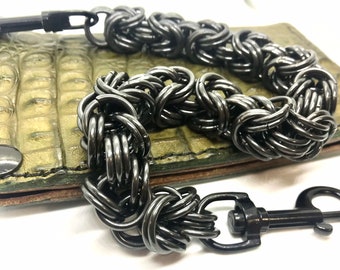 May include: A black metal chain wallet chain with a black metal clasp. The chain is made of multiple interwoven rings. The chain is lying on a green alligator skin wallet.