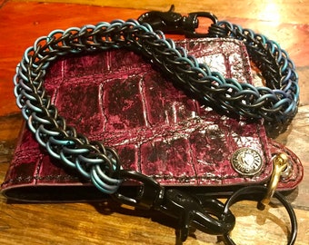 May include: A black and blue chain wallet with a purple alligator skin wallet. The chain has a black and blue braided design. The wallet has a silver snap and a gold ring.