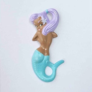 Può includere: Una statuetta di sirena in ceramica con un corpo marrone chiaro, una coda turchese e capelli lavanda. La sirena ha una stella marina nei capelli.