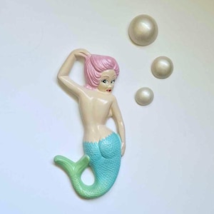 Può includere: Un decoro da parete in ceramica a forma di sirena con uno schema di colori rosa e blu. La sirena si tiene i capelli con una mano e ha una coda verde e blu. Ci sono tre decorazioni bianche a forma di perla intorno alla sirena.