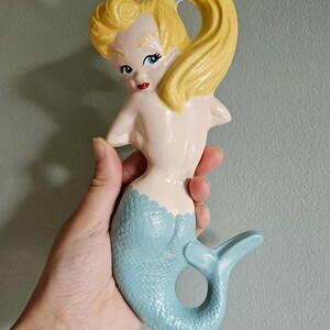 May include: A ceramic mermaid figurine with a light blue tail and a cream-colored body. The mermaid has yellow hair pulled back in a ponytail and red lips. The figurine is held against a light green background.