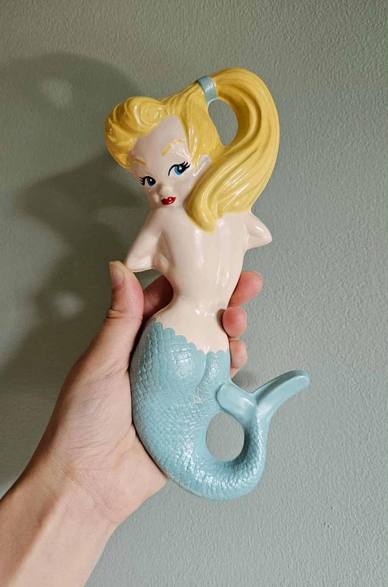 May include: A ceramic mermaid figurine with a light blue tail and a cream-colored body. The mermaid has yellow hair pulled back in a ponytail and red lips. The figurine is held against a light green background.