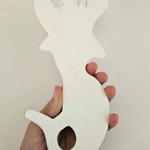 May include: A white, seahorse-shaped wall hanging with a textured surface. The ceramic piece has a hole near the tail and a loop for hanging. The edges are painted with blue and yellow accents.