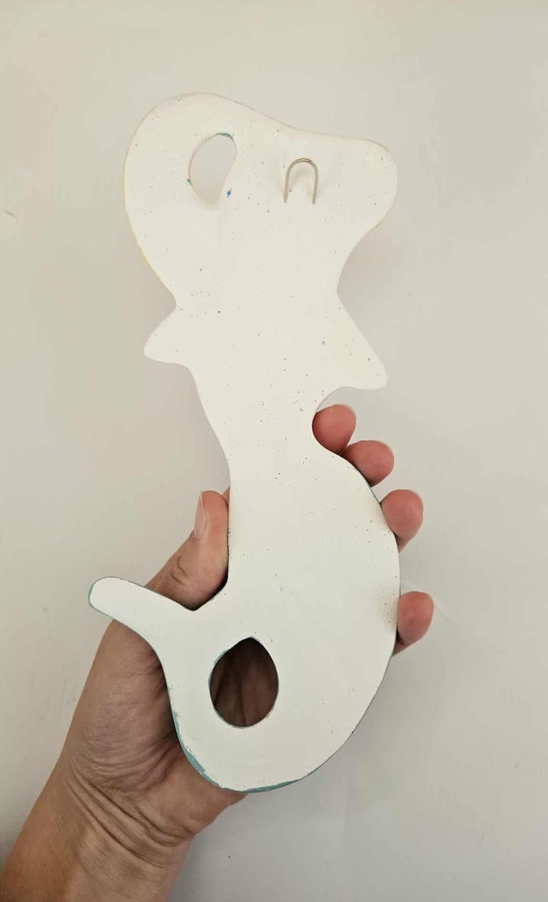 May include: A white, seahorse-shaped wall hanging with a textured surface. The ceramic piece has a hole near the tail and a loop for hanging. The edges are painted with blue and yellow accents.