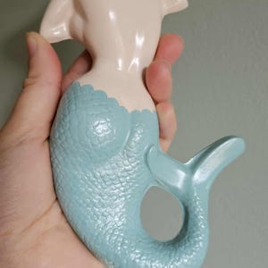 May include: A ceramic mermaid figurine with a light blue tail and a cream-colored body. The mermaid has blonde hair, blue eyes, and red lips. The tail has a textured scale pattern.