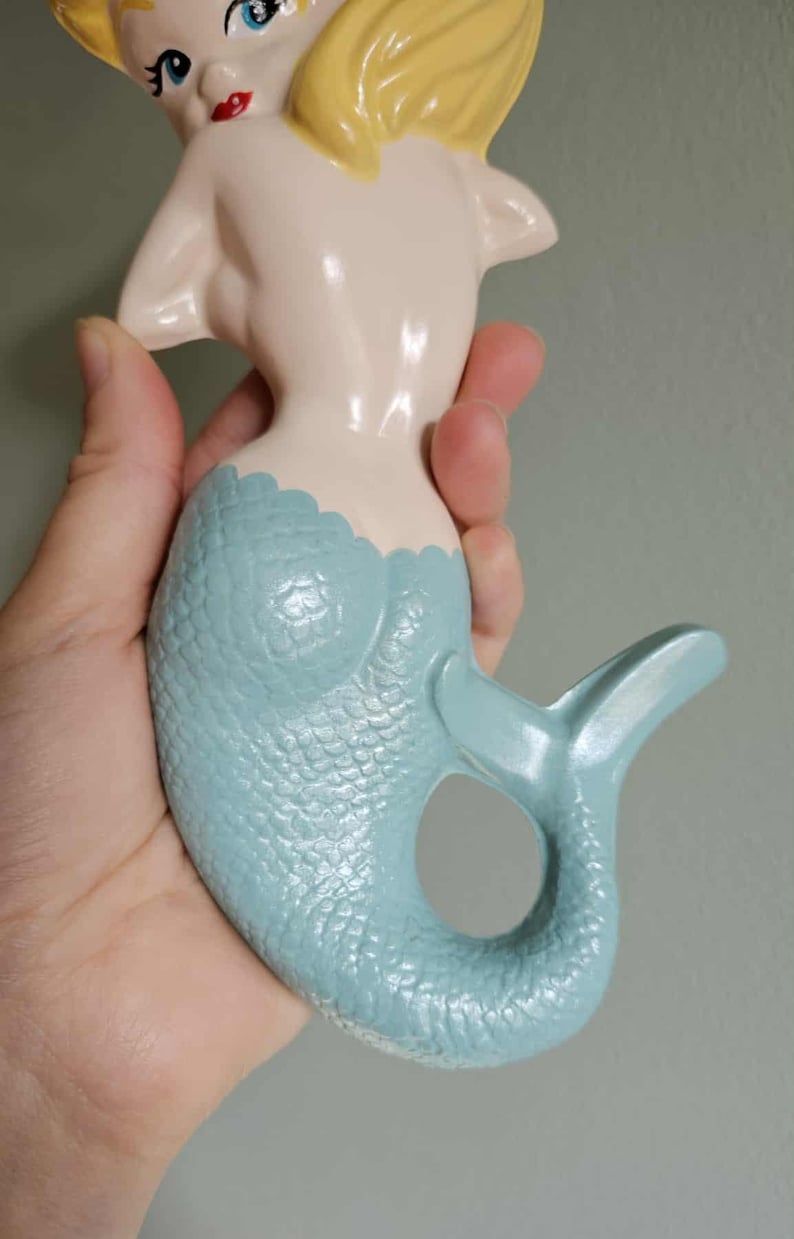 May include: A ceramic mermaid figurine with a light blue tail and a cream-colored body. The mermaid has blonde hair, blue eyes, and red lips. The tail has a textured scale pattern.