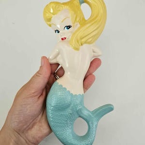 May include: A ceramic mermaid figurine with a light blue tail and blonde hair pulled back in a ponytail. The mermaid has a light skin tone, red lips, and blue eyes. The figurine is held in a hand.