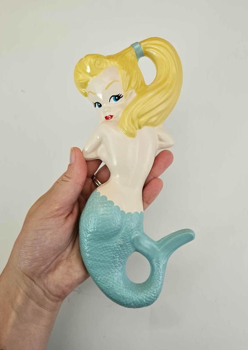 May include: A ceramic mermaid figurine with a light blue tail and blonde hair pulled back in a ponytail. The mermaid has a light skin tone, red lips, and blue eyes. The figurine is held in a hand.
