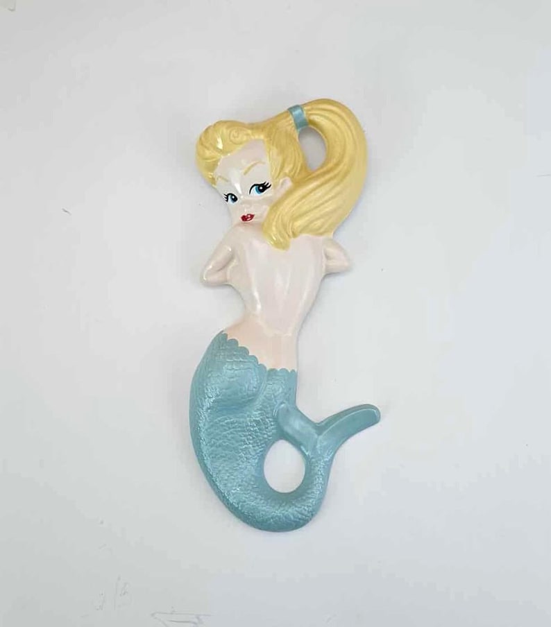 May include: A ceramic mermaid wall hanging. The mermaid has blonde hair tied back with a blue ribbon, blue eyes, and red lips. Her upper body is pale pink, and her tail is light blue. The mermaid is a vintage-style decorative item.