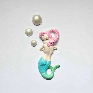 Può includere: Una statuetta di sirena in ceramica con capelli rosa e coda turchese. La sirena è circondata da tre decorazioni bianche a forma di perle.