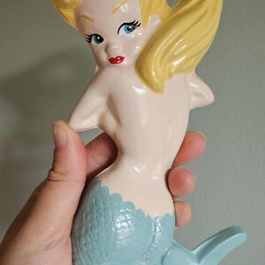 May include: A ceramic mermaid figurine with blonde hair, blue eyes, and red lips. The mermaid's tail is light blue with a textured scale pattern. The figurine is held in a hand.