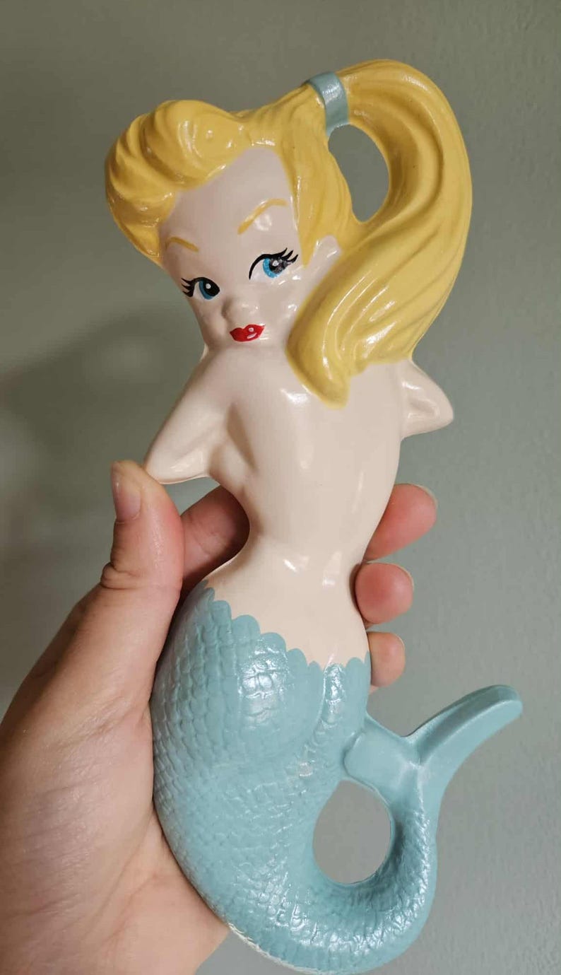 May include: A ceramic mermaid figurine with blonde hair, blue eyes, and red lips. The mermaid's tail is light blue with a textured scale pattern. The figurine is held in a hand.