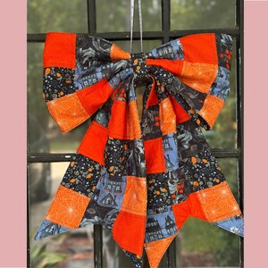 May include: A large decorative bow made of fabric featuring a patchwork design. The pattern includes solid orange squares, orange spiderwebs on black, and illustrations of dark houses and trees on a blue background. It hangs from a thin white string.