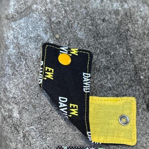 May include: A black fabric cord keeper featuring a repeating yellow text pattern that reads "EW, DAVID". The accessory includes a bright yellow snap button on the exterior and a metallic snap fastener on the interior yellow fabric lining.