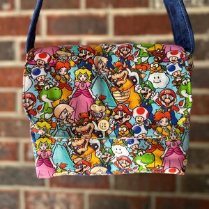 May include: A rectangular shoulder bag with a flap closure, featuring a colorful print of various characters from the Super Mario video game series. The bag has a blue strap and a light-colored button closure. The background is a brick wall.