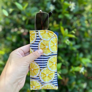 May include: A rectangular fabric glasses case featuring a pattern of yellow lemon slices over a navy blue and white horizontal striped background. The case is quilted with visible yellow stitching and contains a pair of dark-framed sunglasses.