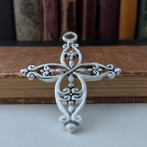Peut inclure: Un pendentif en forme de croix en filigrane argenté avec des détails complexes. La croix repose sur un livre relié en cuir brun.