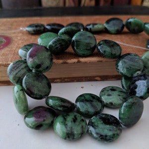 May include: A strand of green and black gemstone beads, with a slightly oval shape. The beads are strung on a clear thread and are arranged in a loose circle.