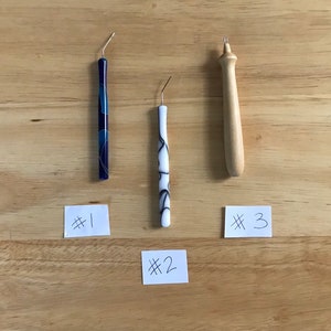 May include: Three dental tools with curved metal tips are displayed on a wooden surface. One tool has a blue and white patterned handle, another has a white handle with black swirls, and the third is made of light-colored wood. Each tool is labeled with a number.