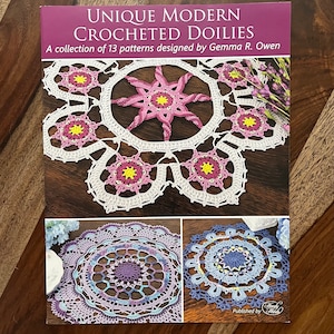 May include: Book cover featuring a collection of 13 crochet doily patterns. The cover shows three doilies in white, purple, and blue. The title is "Unique Modern Crocheted Doilies" and the book is published by Handy Stitch.