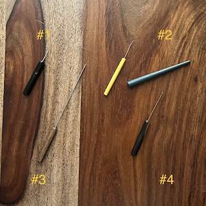 May include: Four different types of crochet hooks, numbered 1 through 4, are laid out on a wooden surface. The hooks are made of metal and wood and are different colors, including black, yellow, and brown.