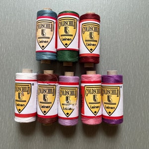 May include: Ten spools of thread in various colors, including blue, green, red, brown, pink, and purple. Each spool has the word "Goldschild" printed on it, along with the word "Leinen", which is German for linen.