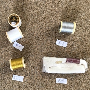 May include: Four spools of thread, two white, one gold, and one silver. The spools are labeled with numbers 1 through 4.