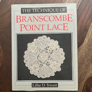 May include: A book titled "THE TECHNIQUE OF BRANSCOMBE POINT LACE" by Lillie D. Trivett. The cover features a detailed, circular lace design in white against a black background, framed by a gray border. The book is on a wooden surface.