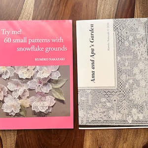 Peut inclure: Deux livres posés sur une surface en bois. Le livre de gauche a une couverture rose avec le texte "Try me! 60 small patterns with snowflake grounds" et des illustrations florales. Le livre de droite a le texte "Ama and Apa's Garden".