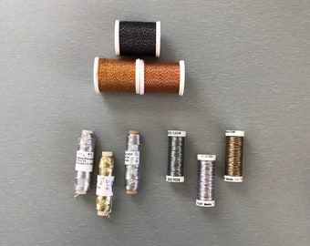 May include: Assortment of metallic embroidery floss spools in various colors, including gold, silver, copper, and black. The spools are arranged in a pyramid shape on a gray surface.