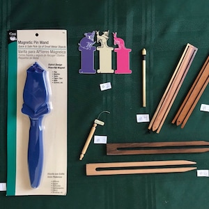 May include: A blue plastic magnetic pin wand with a rose design. The wand is packaged on a white card with the text "Magnetic Pin Wand" and "Quick & Safe Pick-Up of Small Metal Objects". The image also shows various wooden tools for needlework, including a wooden needle threader, a wooden needle minder, and a wooden knitting needle.