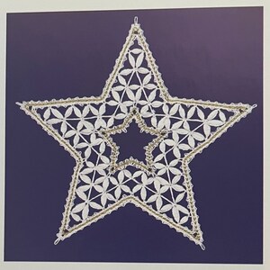May include: A white, star-shaped, handmade lace ornament with a gold-colored border and a dark blue background. The star is filled with a floral pattern and has a smaller star shape in the center. The ornament is likely for holiday or home decor.