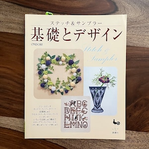 May include: A book titled "Stitch & Sampler: Basics and Design" by Ondori. The cover features a wreath of flowers and berries, a vase with flowers, and a sampler with the alphabet. The book is published by Yokai.