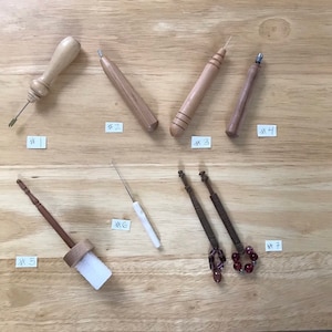 May include: Assortment of seven wooden tools, likely for crafts or needlework, arranged on a light brown wooden surface. The tools vary in shape and size, with some featuring metal tips and others with decorative elements. Each tool is labeled with a number.