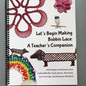 May include: A book titled "Let's Begin Making Bobbin Lace: A Teacher's Companion" with design contributions from Cathy Belleville, Paula Harten, Kim Davis, Jane Partridge, and Rosemary Shepherd. The cover features various bobbin lace designs, including a flower, a bird, a dachshund, and a rainbow.