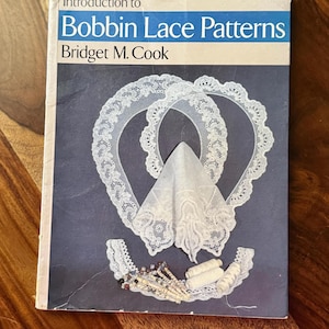 May include: A book titled "Introduction to Bobbin Lace Patterns" by Bridget M. Cook. The cover features a blue background with white lace designs and a white handkerchief. The book is on a wooden surface.