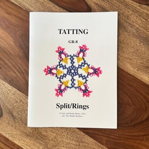 May include: A white booklet titled "TATTING GR-8 Split/Rings" with a colorful, intricate design in the center. The design features a blue, yellow, and pink floral pattern. The booklet is on a wooden surface.