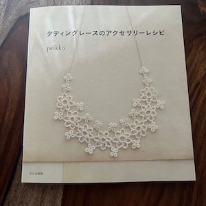 May include: A book titled "タティングレースのアクセサリーレシピ" by peikko, featuring a white lace necklace. The necklace is made of delicate, flower-shaped lace elements. The book cover is light beige, and the necklace is displayed on the cover.