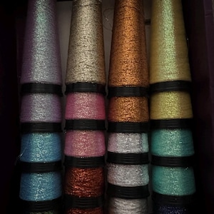 May include: A collection of sixteen spools of metallic thread arranged in four vertical columns. The spools feature a variety of shimmering colours including lavender, gold, copper, yellow, blue, pink, silver, green, and dark purple, each separated by black rings.