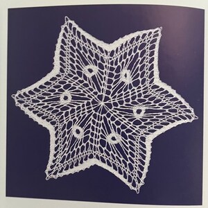 May include: A white, star-shaped doily with a delicate, openwork design. The doily features a six-pointed star shape with a central pattern and circular accents. The intricate lace-like pattern is set against a dark blue background.
