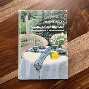 Peut inclure: Un livre intitulé "Nordische Tischbänder" d'Ursula Städtke. La couverture du livre présente une nappe blanche avec un chemin de table en crochet bleu et noir, deux chaises en osier blanches et une rose jaune sur la table. Le livre est publié par Barbara Fay Verlag.