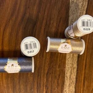 May include: Four spools of DMC Diamant embroidery floss. Two spools are gold, one is silver, and one is dark gray. Each spool has a label with the DMC logo, the words "Diamant" and "Art. 380", and a barcode. The labels also include the DMC color numbers: D225, D317, and D317.