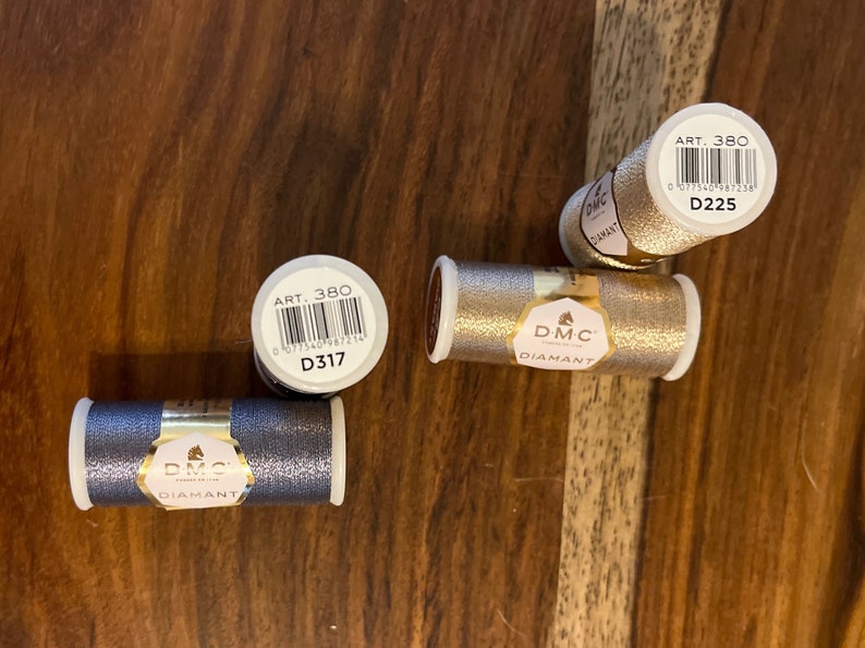 May include: Four spools of DMC Diamant embroidery floss. Two spools are gold, one is silver, and one is dark gray. Each spool has a label with the DMC logo, the words "Diamant" and "Art. 380", and a barcode. The labels also include the DMC color numbers: D225, D317, and D317.