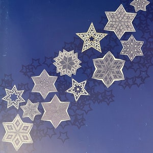 May include: A collection of white, intricately designed star ornaments arranged on a dark blue background with star-shaped shadows. The stars vary in size and pattern, creating a decorative display. The ornaments appear to be made of lace or a similar delicate material.