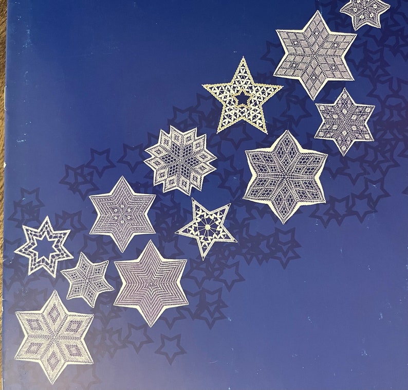 May include: A collection of white, intricately designed star ornaments arranged on a dark blue background with star-shaped shadows. The stars vary in size and pattern, creating a decorative display. The ornaments appear to be made of lace or a similar delicate material.