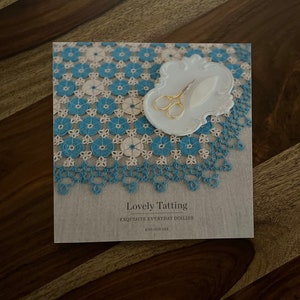 May include: A book titled "Lovely Tatting: Exquisite Everyday Doilies" by Hye-Oon Lee. The cover features a close-up of a blue and white crocheted doily with a pair of gold scissors and a white ceramic needle holder.