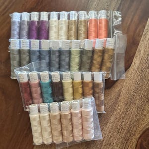 May include: A collection of spools of silk thread in various colors, including shades of purple, green, orange, and pink. The spools are packaged in clear plastic and are labeled with text. The threads are likely for sewing or embroidery projects.