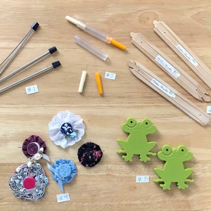 May include: An assortment of craft supplies, including ribbon threaders, needles, flower embellishments, and green frog-shaped cutouts. The items are arranged on a light wood-grain surface. The image is well-lit and shows the details of each item.