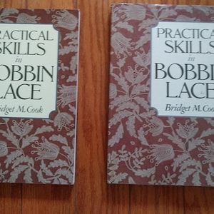 May include: Two brown books with a white floral design on the cover. The title of the books is "Practical Skills in Bobbin Lace" by Bridget M. Cook.